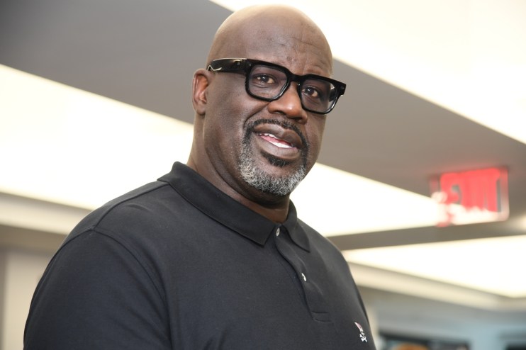 Shaquille ONeal in a black polo shirt and black-framed glasses, smiling slightly, with a blurred background.