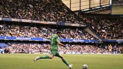 Goalkeeper Emiliano Martinez in green uniform kicking a soccer ball on a field in a packed stadium.