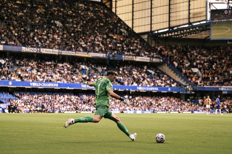Goalkeeper Emiliano Martinez in green uniform kicking a soccer ball on a field in a packed stadium.