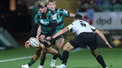 Rugby player in green and black uniform passing a Gilbert ball during a match, opponent in white jersey close by.
