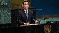 Man in suit and glasses speaking at a podium with the UN emblem, microphones, and a clear screen.