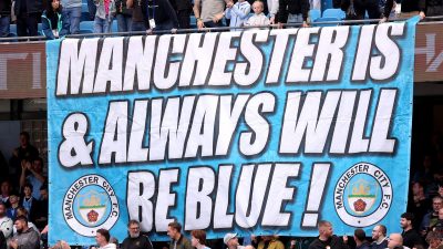 A large banner reads Manchester Is & Always Will Be Blue! with Manchester City F.C. crests, surrounded by cheering fans.