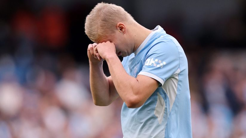 Erling Haaland, a blonde male footballer in a light blue jersey, rubs his eyes in frustration on the field.