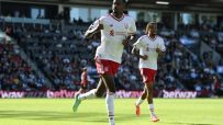 Two Liverpool FC players in white and red kits run on a sunny green football pitch during a match.