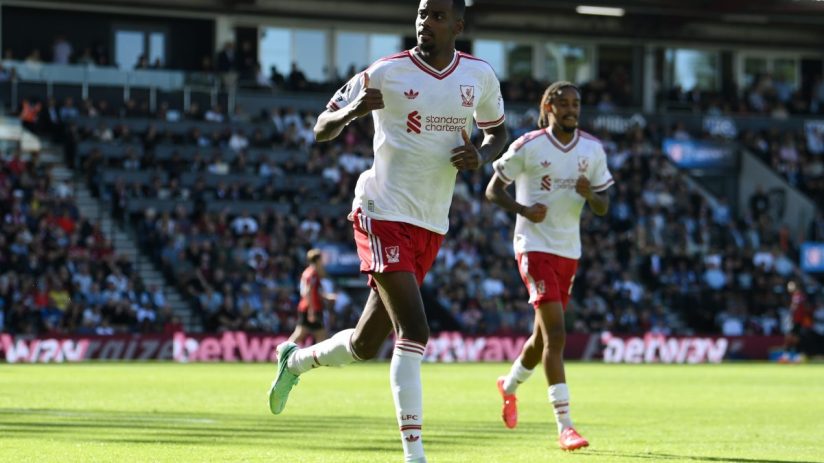 Two Liverpool FC players in white and red kits run on a sunny green football pitch during a match.