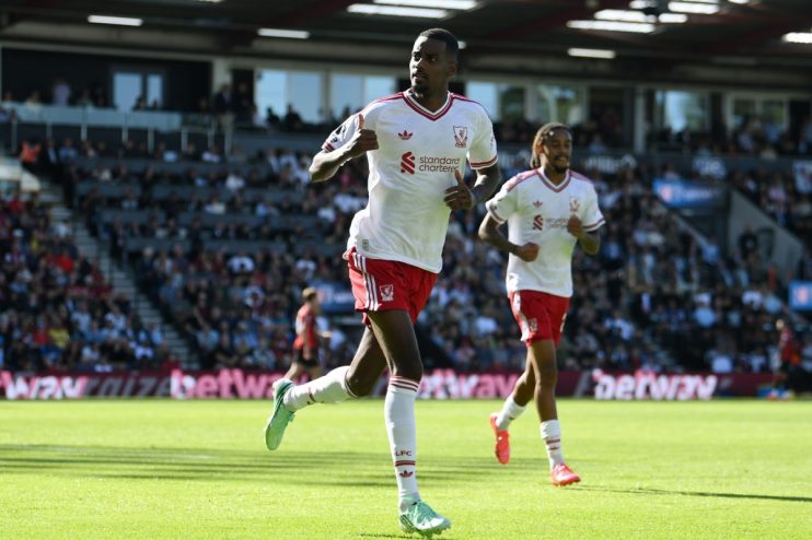 Two Liverpool FC players in white and red kits run on a sunny green football pitch during a match.