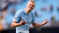 Erling Haaland in a Manchester City kit, smiling with arms outstretched on a bright day.