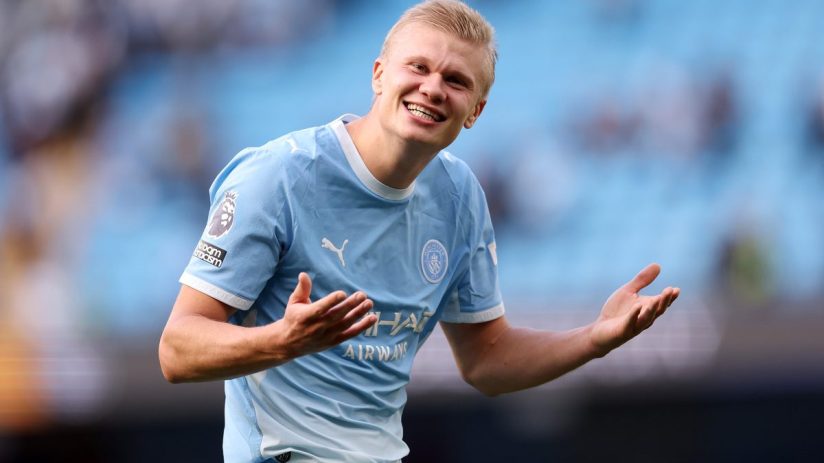 Erling Haaland in a Manchester City kit, smiling with arms outstretched on a bright day.