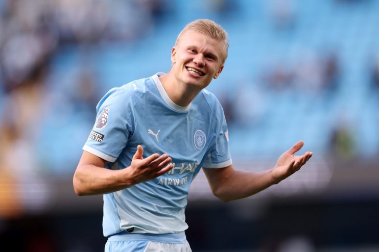 Erling Haaland in a Manchester City kit, smiling with arms outstretched on a bright day.