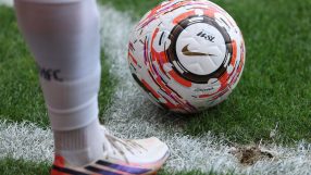 Football players leg and foot in white kit next to a Nike WSL football on green grass with a white line.