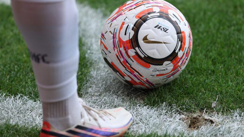 Football players leg and foot in white kit next to a Nike WSL football on green grass with a white line.