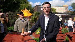Andy Burnham, Mayor of Greater Manchester, painting a red picnic table outdoors with a brush.