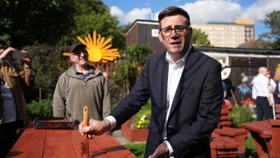 Andy Burnham, Mayor of Greater Manchester, painting a red picnic table outdoors with a brush.