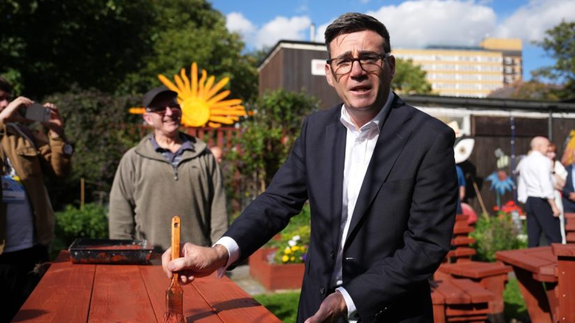Andy Burnham, Mayor of Greater Manchester, painting a red picnic table outdoors with a brush.