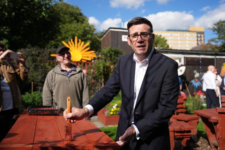 Andy Burnham, Mayor of Greater Manchester, painting a red picnic table outdoors with a brush.