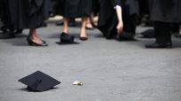Black graduation cap with tassel on asphalt, feet of graduates in robes in background.