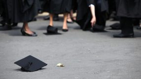 Black graduation cap with tassel on asphalt, feet of graduates in robes in background.