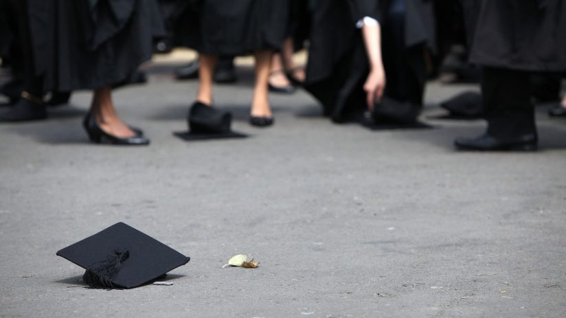 Black graduation cap with tassel on asphalt, feet of graduates in robes in background.