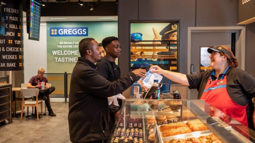 Greggs employee serving two customers at the counter in a Gatwick Airport store, with baked goods on display.