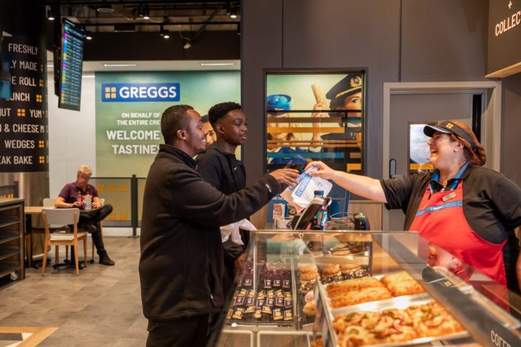 Greggs employee serving two customers at the counter in a Gatwick Airport store, with baked goods on display.