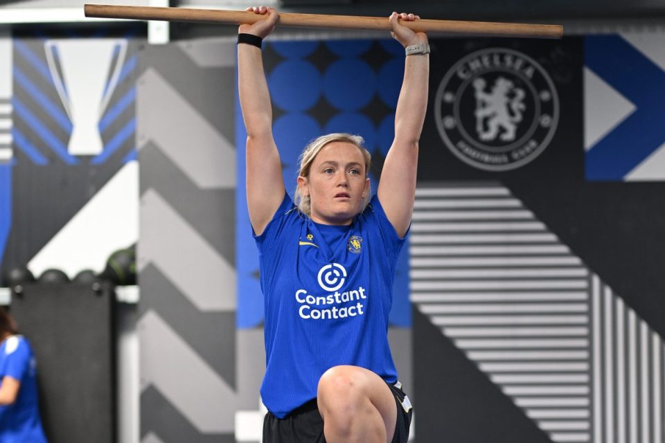 Female athlete in blue uniform with Constant Contact logo, balancing on one leg, holding a wooden bar overhead.