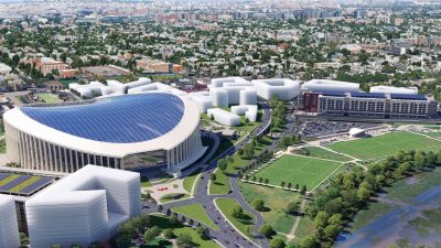Aerial view of a modern stadium with a solar panel roof, surrounded by new development, green fields, and a city