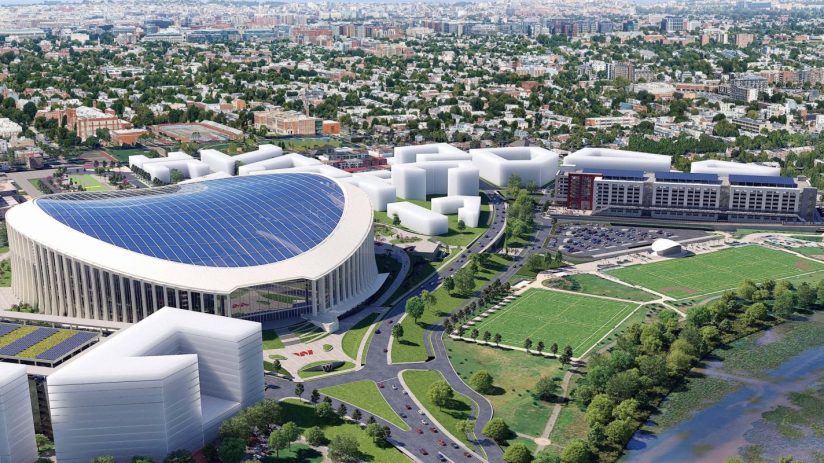 Aerial view of a modern stadium with a solar panel roof, surrounded by new development, green fields, and a city