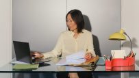 Professional woman working on laptop at a glass desk, reviewing documents in an office setting