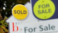Real estate signs: a yellow SOLD sign and a blurred green FOR SALE sign, indicating house prices and market activity.