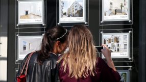 Two women view London housing property listings in a real estate agents window, one takes a phone photo.