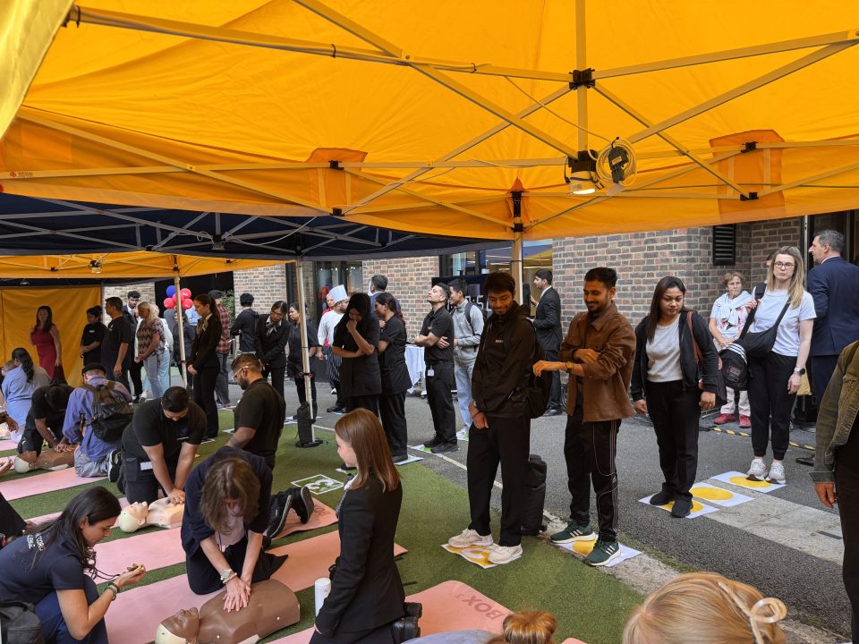 People practicing CPR on mannequins and observing under a yellow tent at a public event.