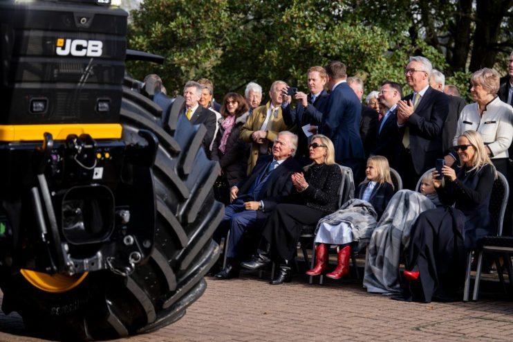 Audience observing a JCB machine, with a large tire prominent in the foreground.