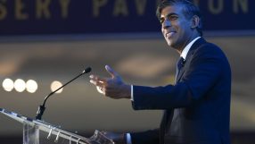Rishi Sunak speaking at a podium, gesturing with his hand, at an event with a blurred sign in background.