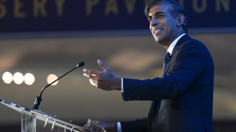 Rishi Sunak speaking at a podium, gesturing with his hand, at an event with a blurred sign in background.