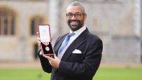 James Cleverly, in a suit and tie, smiling and holding an open red box with a medal, outdoors with buildings in background.