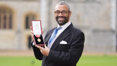 James Cleverly, in a suit and tie, smiling and holding an open red box with a medal, outdoors with buildings in background.
