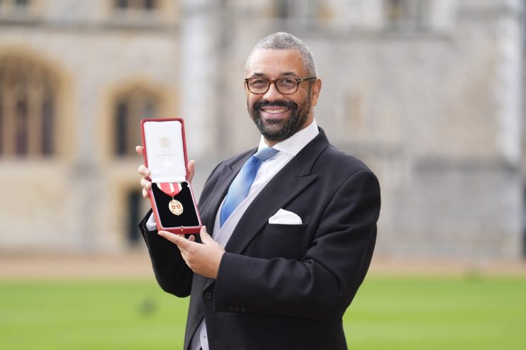 James Cleverly, in a suit and tie, smiling and holding an open red box with a medal, outdoors with buildings in background.
