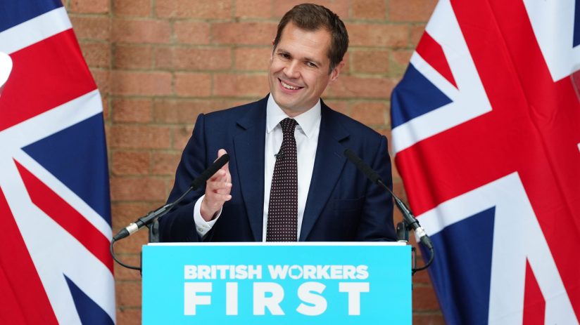 Robert Jenrick speaking at a podium with British Workers First and Union Jack flags, discussing bank taxes.