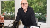 John Caudwell, bald man in black shirt and glasses, smiling, leaning on a desk with window garden background.