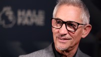 Gary Lineker smiling in black-rimmed glasses, wearing a gray suit jacket, blurred Hall of Fame in background