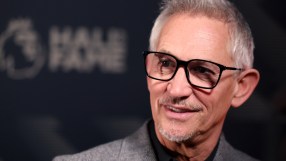 Gary Lineker smiling in black-rimmed glasses, wearing a gray suit jacket, blurred Hall of Fame in background