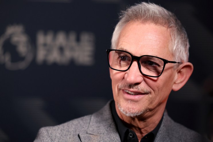 Gary Lineker smiling in black-rimmed glasses, wearing a gray suit jacket, blurred Hall of Fame in background