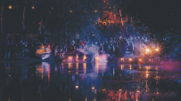 Lost Village festival at night: crowd gathered by a lake, lit by lanterns and colorful lights, reflecting on the water.