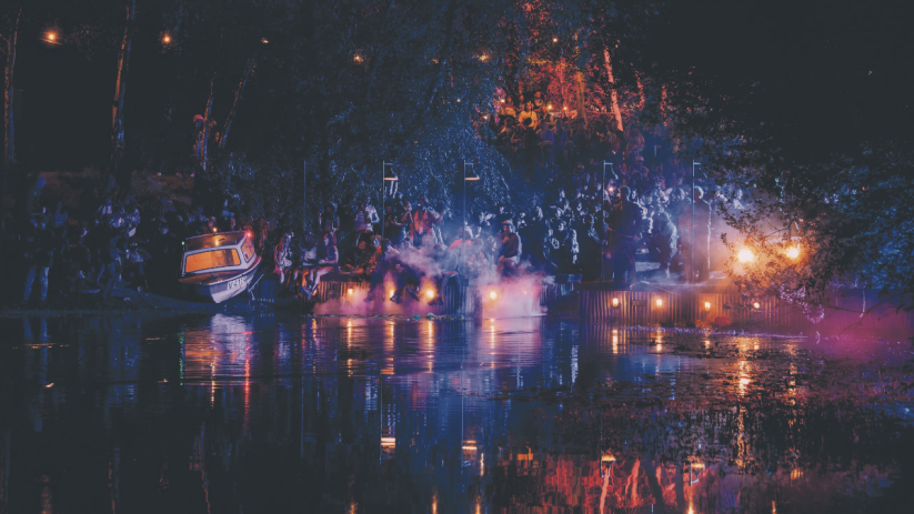 Lost Village festival at night: crowd gathered by a lake, lit by lanterns and colorful lights, reflecting on the water.