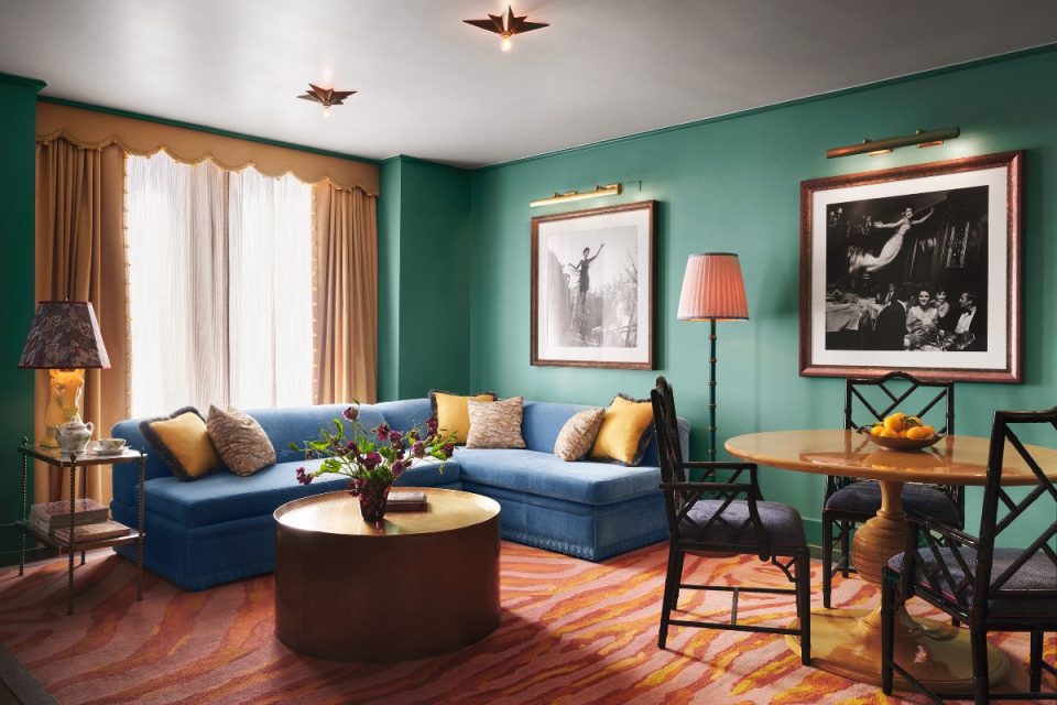 Luxurious suite at The Langham New York hotel with teal walls, blue sectional sofa, gold coffee table, and dining area.