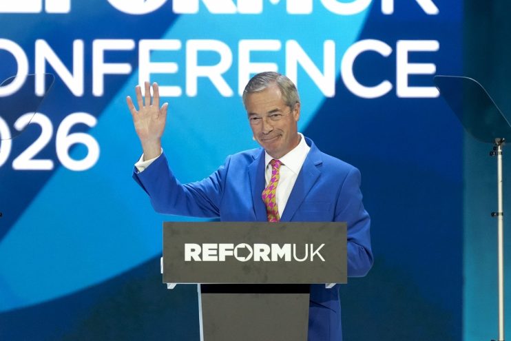 Nigel Farage at a Reform UK conference, gesturing from a podium with the party logo and CONFERENCE 2026 in background