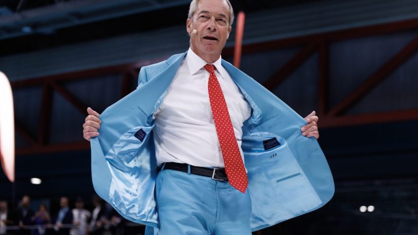 Nigel Farage in a light blue suit and red polka-dot tie, speaking at the Reform conference