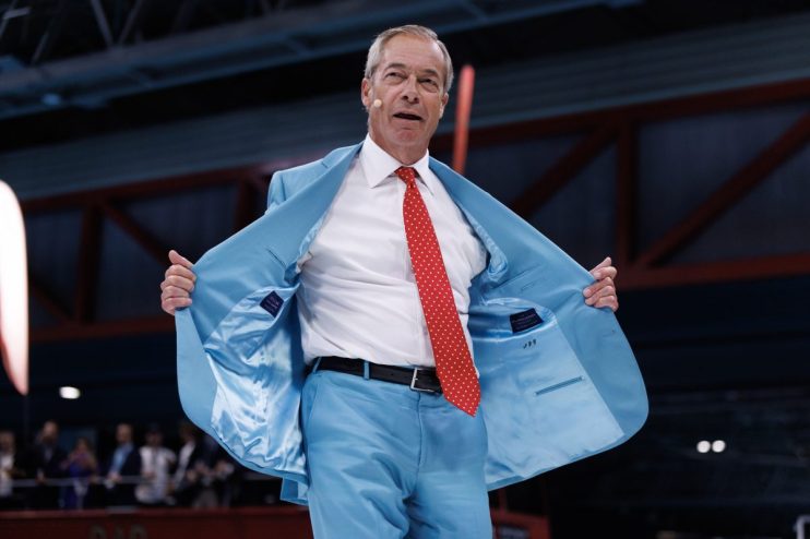 Nigel Farage in a light blue suit and red polka-dot tie, speaking at the Reform conference