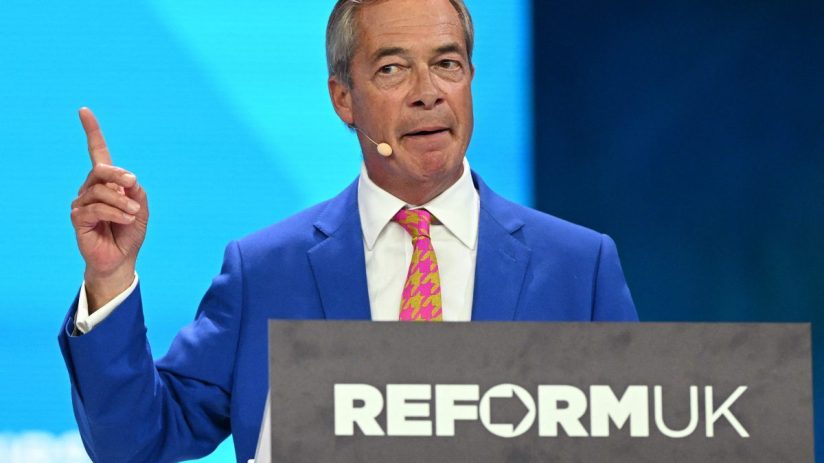 Nigel Farage speaking at a Reform UK event, wearing a blue suit and pink patterned tie, pointing with his right hand.