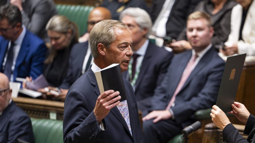 Nigel Farage in Parliament, addressing a female clerk holding a document, with other parliamentarians in the background.